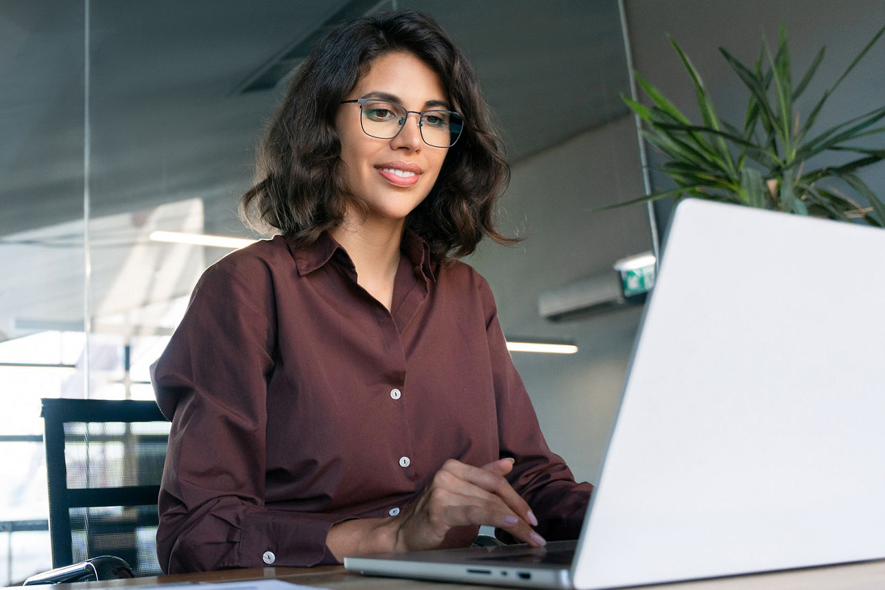 Frau sitzt am Laptop an einem Schreibtisch vor einer Glaswand mit Blick auf Büroräume