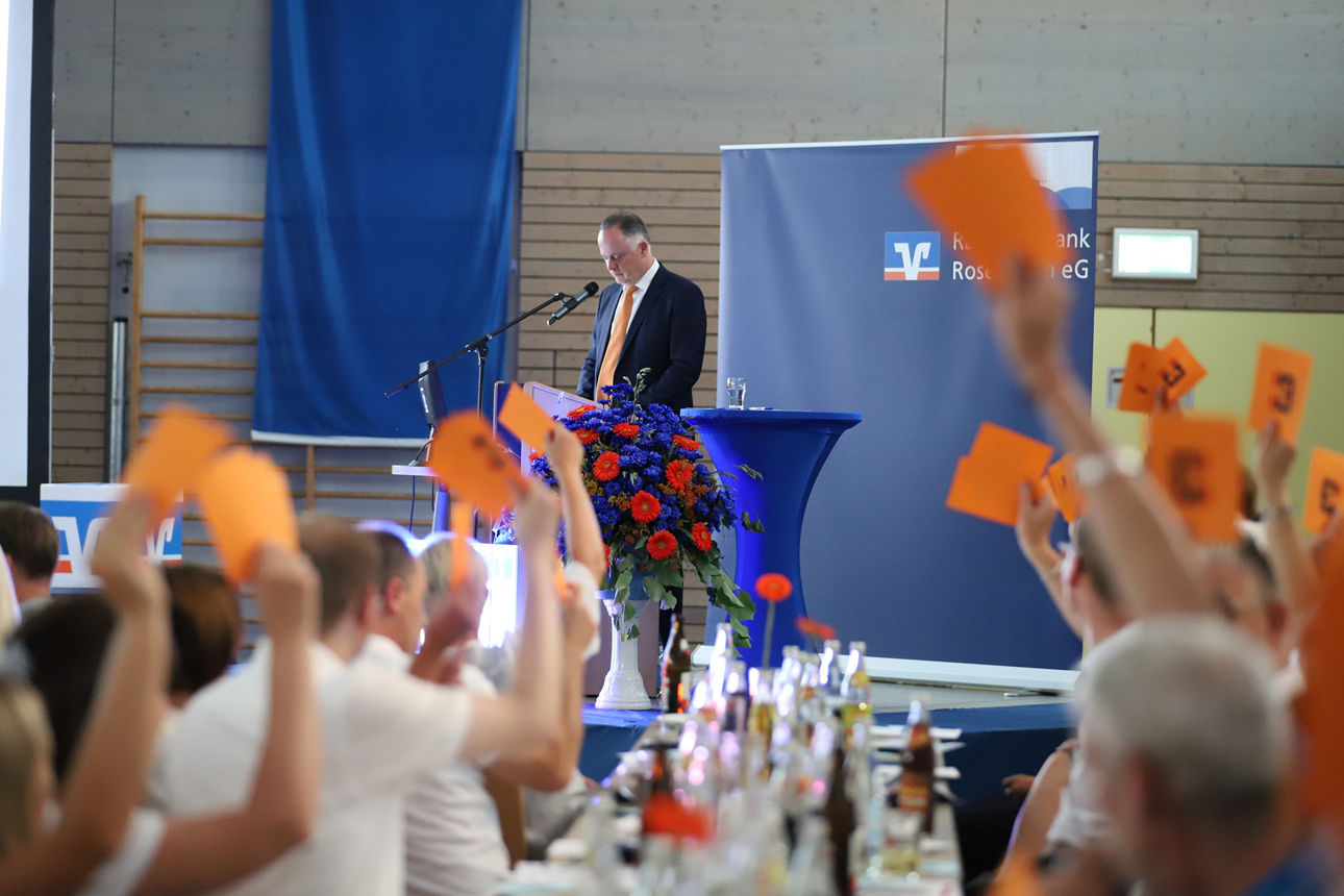 Ein Redner am Blumenpult, während im Vordergrund orange Stimmkarten gehoben werden.