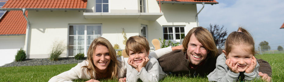 Eine vierköpfige Familie liegt im Gras vor ihrem Haus und lächelt in die Kamera.