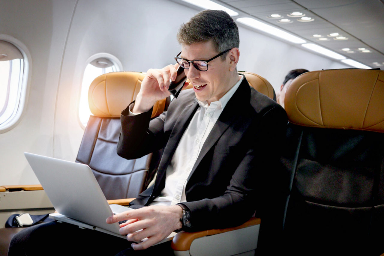 Mann im Flugzeug sitzend mit Handy am Ohr und aufgeklapptem Laptop auf dem Schoß