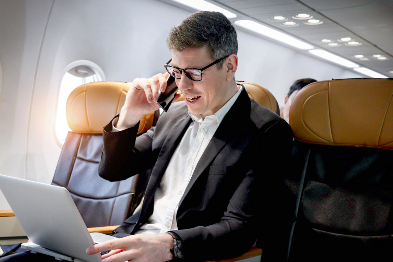 Mann im Flugzeug sitzend mit Handy am Ohr und aufgeklapptem Laptop auf dem Schoß