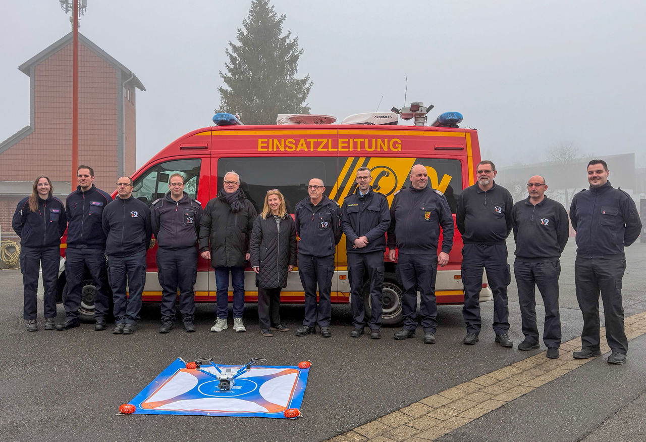 Im Vordergrund fliegt eine BOS-Drohne vom Typ M4TD. Im Hintergrund stehen Feuerwehrleute vor einem Rettungswagen.