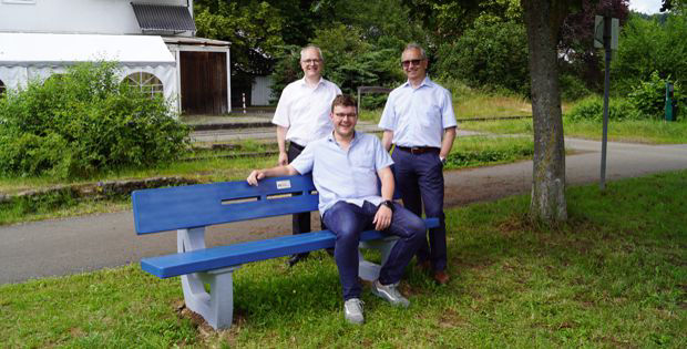 Drei Männer stehen neben einer blauen Parkbank.