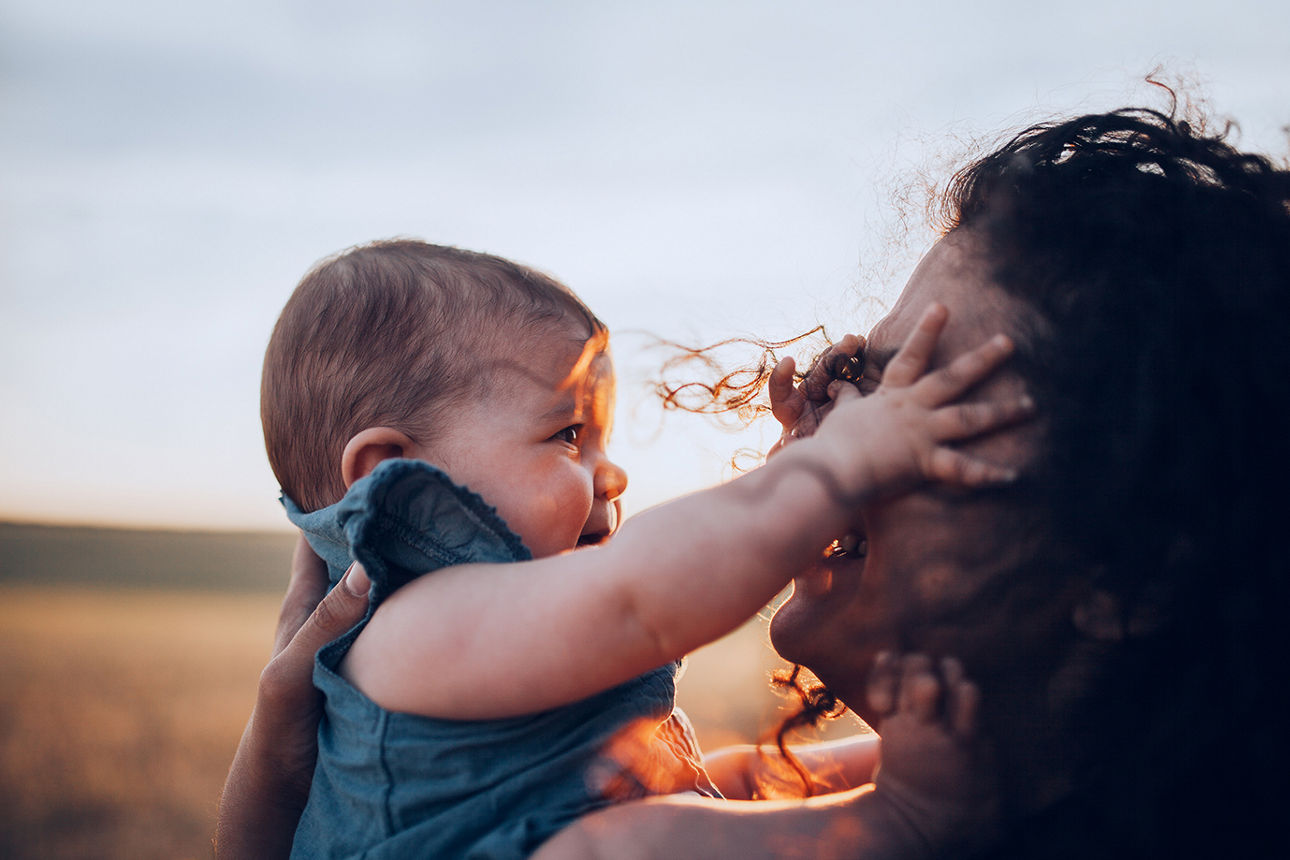 Frau hält ihr Baby im Freien, beide schauen sich an und lächeln