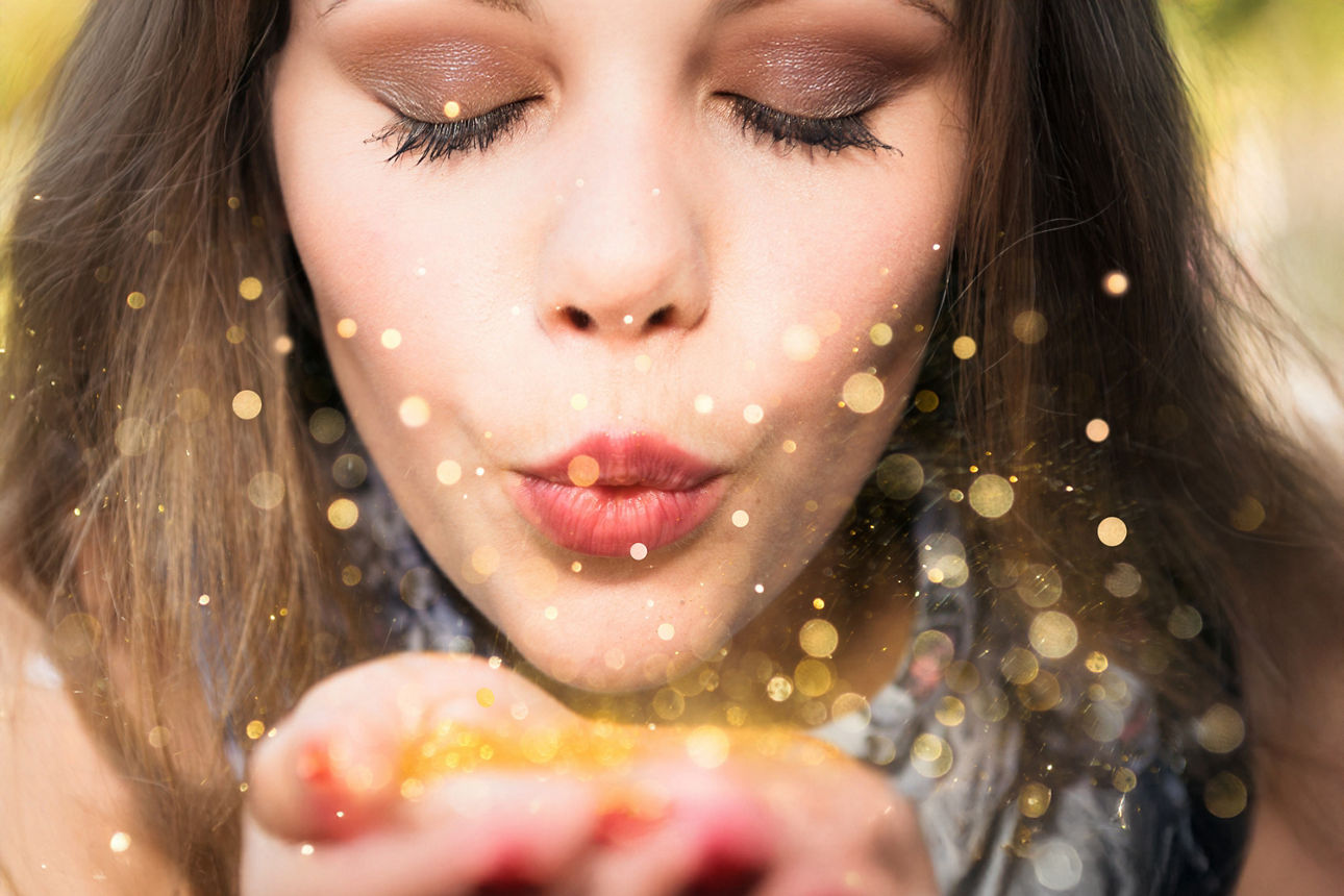 Eine junge Frau pustet funkelnde goldene Partikel aus ihren Händen und erzeugt einen magischen, lichtvollen Moment.