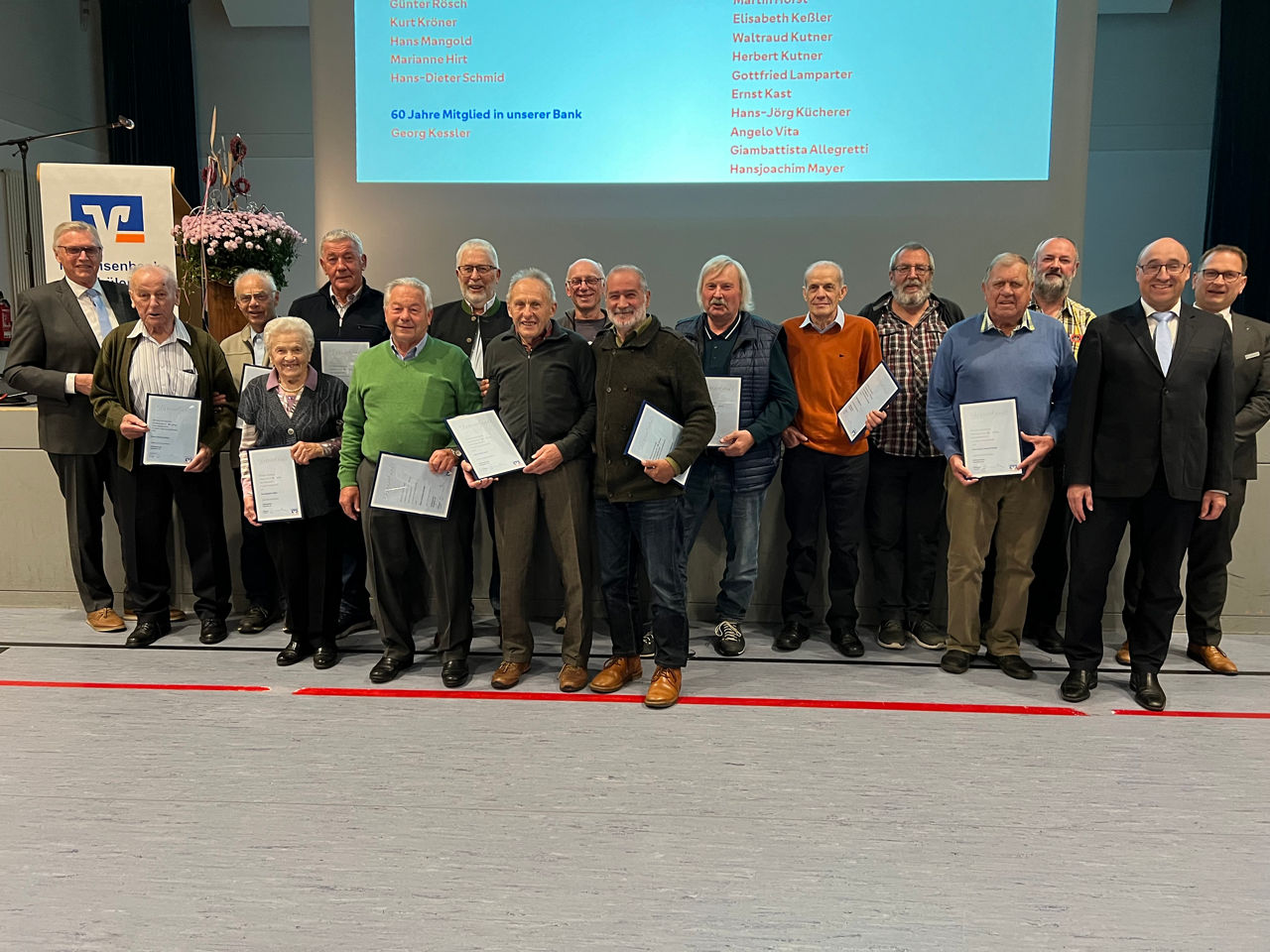 Eine Gruppe von Männern steht vor einer Bühne mit einem großen Bildschirm, auf dem eine Liste von Namen und der Text "60 Jahr