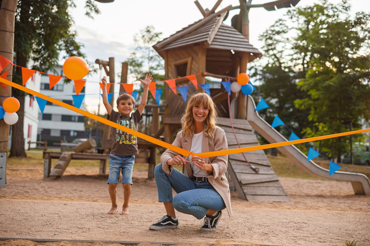 Frau schneidet Band auf Kinderspielplatz durch während sich im Hintergrund ein Kind freut