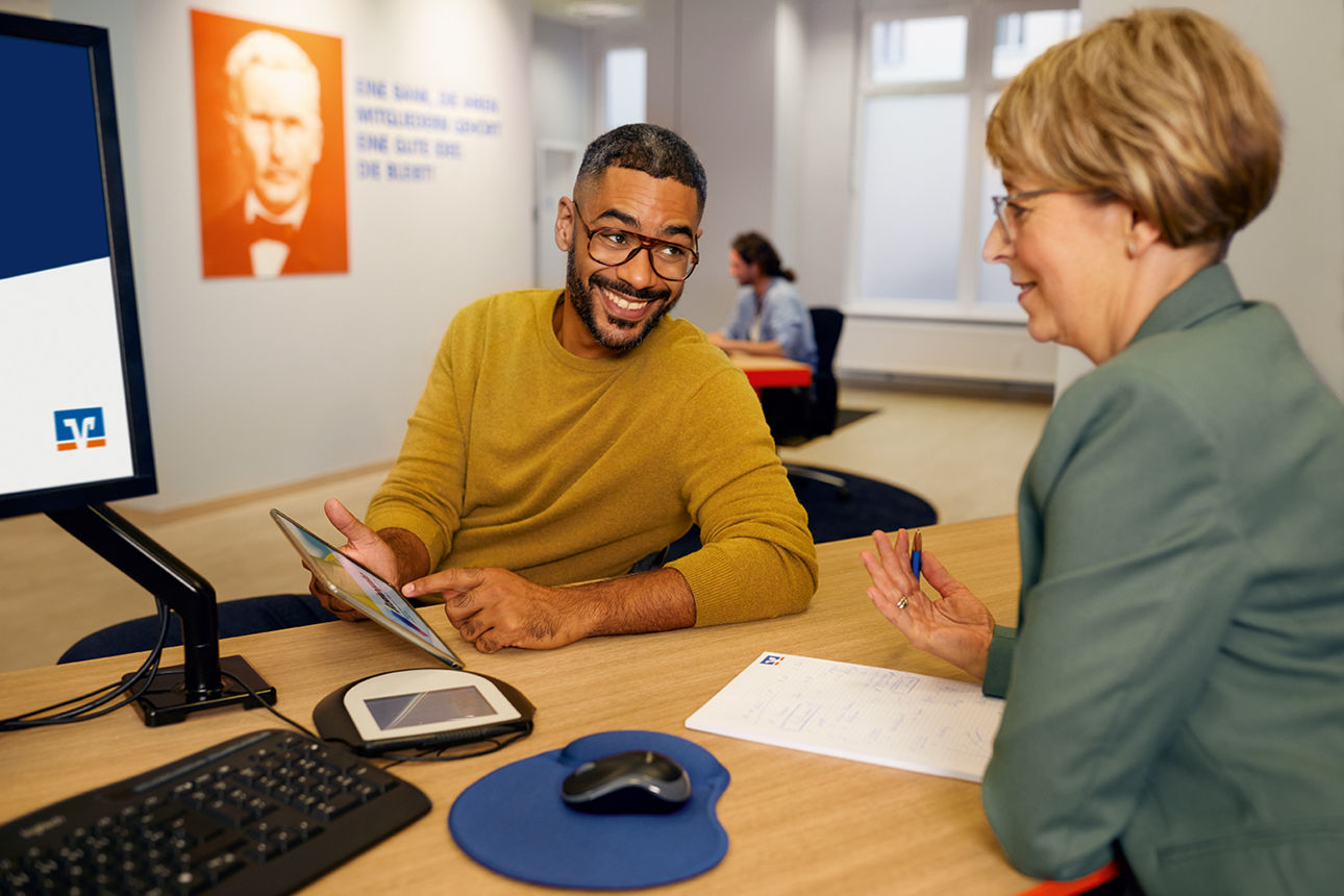 Zwei Personen sitzen an einem Schreibtisch, eine hält ein Tablet, auf dem Tisch liegen ein Computermauspad, eine Maus und ein