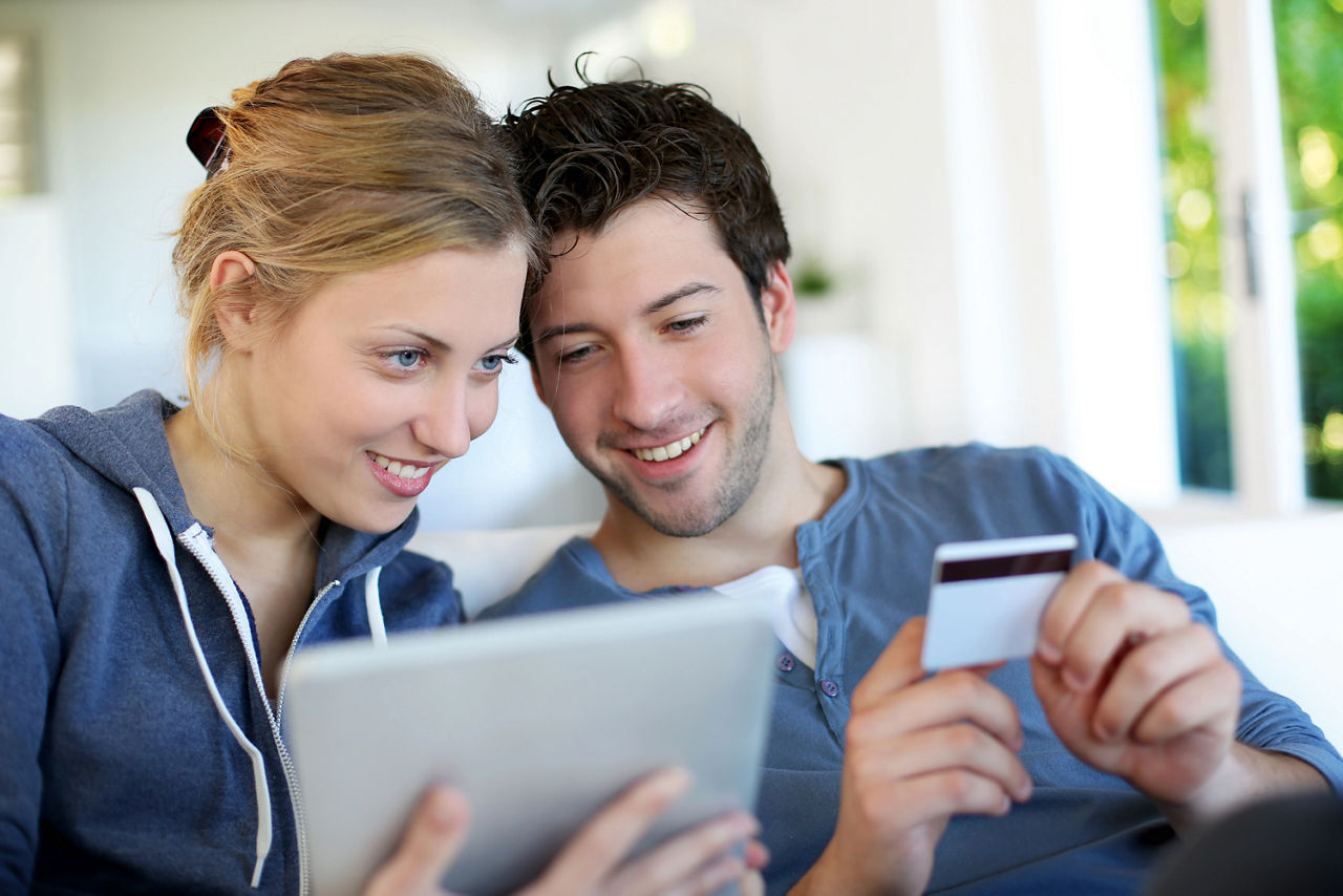 Mann mit einer girocard in der Hand und eine Frau mit einem Tablet in der Hand sitzen zusammen auf einem Sofa 