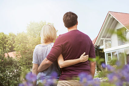Paar hat sich im Arm und blickt auf ein Haus im Hintergrund, sie stehen mit dem Rücken zur Kamera