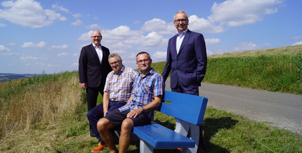 Four men stand and sit on a blue bench in a rural setting.