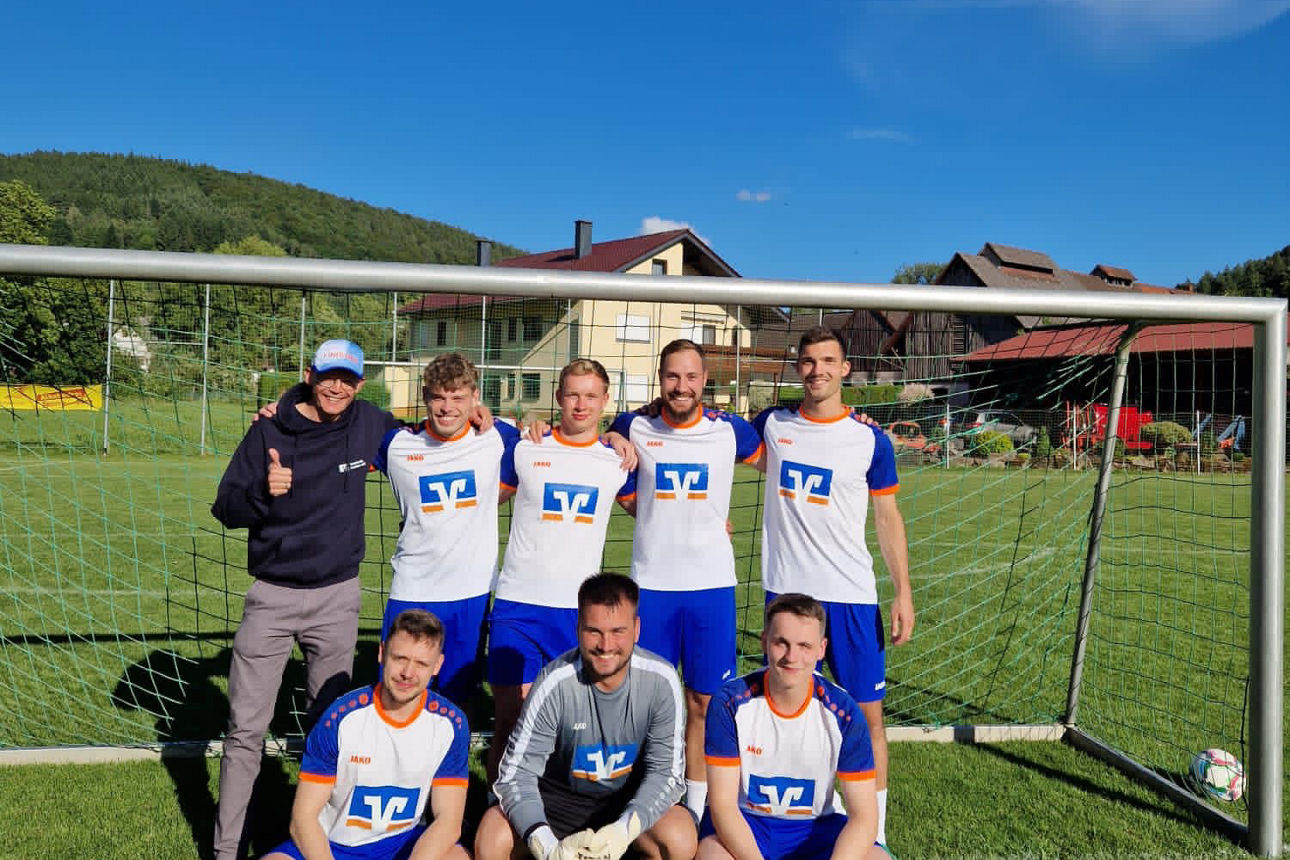 Sieben Spieler und ein Fan posieren vor dem Tor; klarer blauer Himmel und Dorfhäuser im Hintergrund.