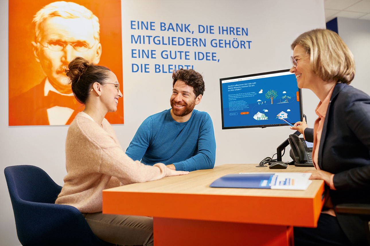 Juges Paar sitzt Beraterin an einem orangefarbenen Tisch, die Beraterin zeigt auf einen Bildschirm mit Grafiken und Text