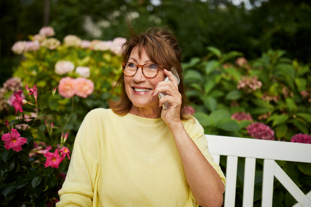 Frau telefoniert auf einer Bank im Garten und lächelt dabei.