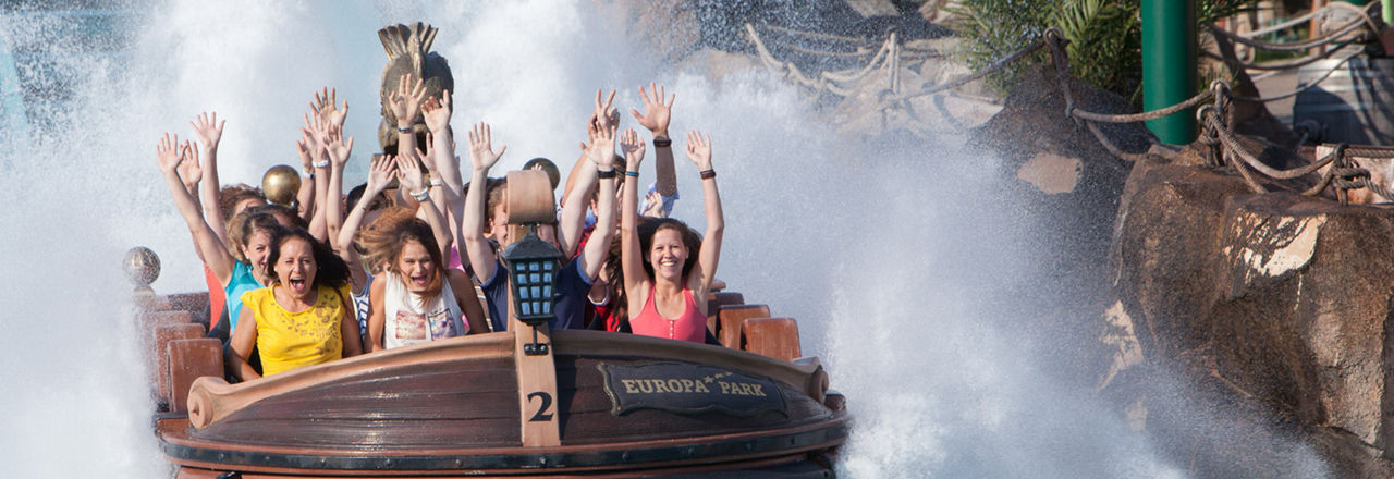 Viele Personen, davon einige Kinder, sitzen in einem Wasser-Fahrgeschäft im Europapark und heben die Hände in die Höhe. Links und rechts spritzt Wasser nach oben.
