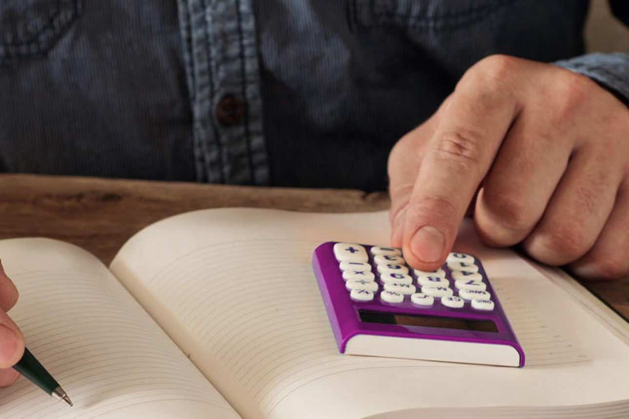 Eine Person, die an einem Tisch sitzt, einen Taschenrechner benutzt und in ein Notizbuch schreibt.