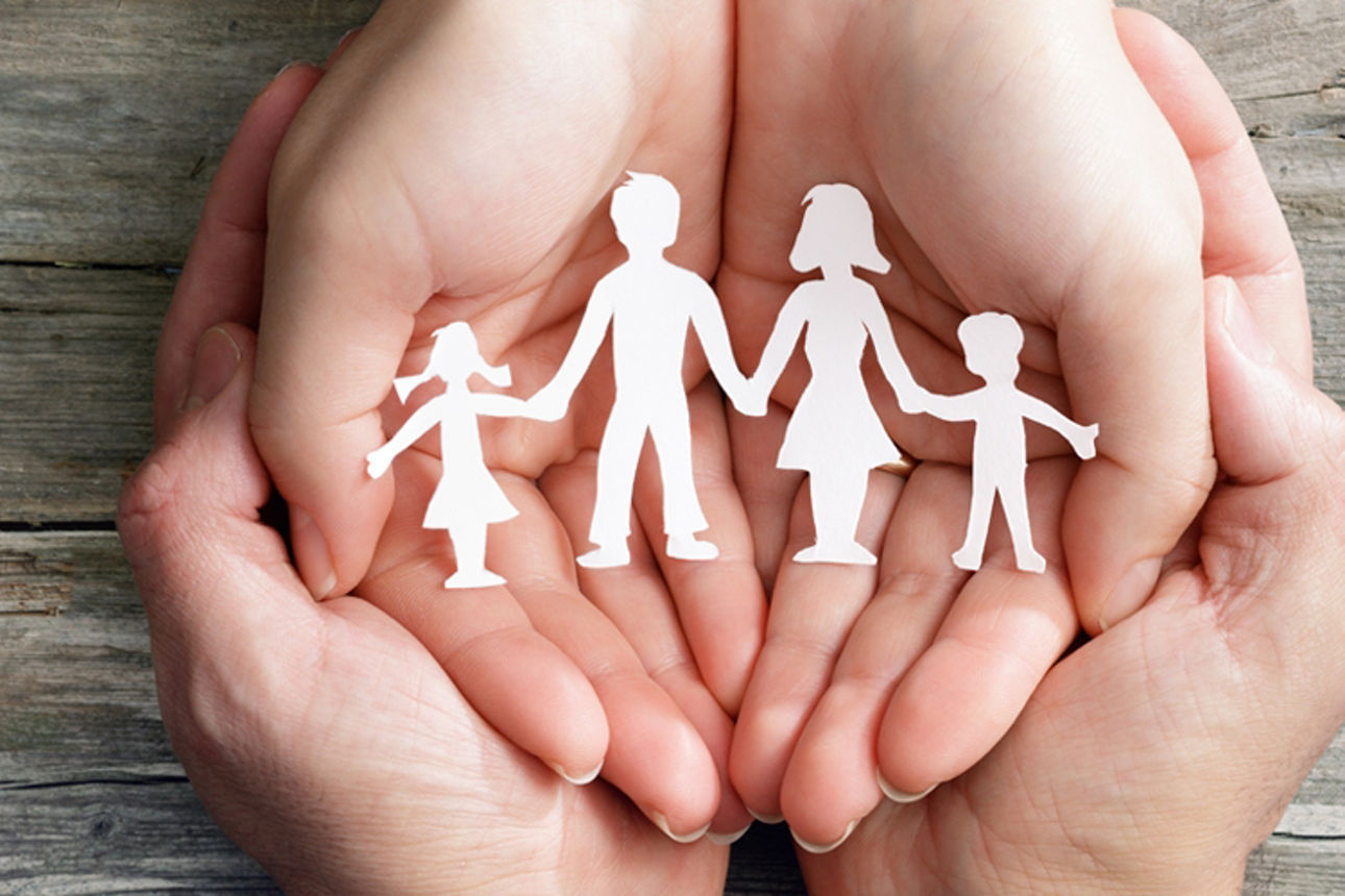 Zwei Personen halten die Hände schützend darin ein Scherenschnitt einer Familie mit Kindern