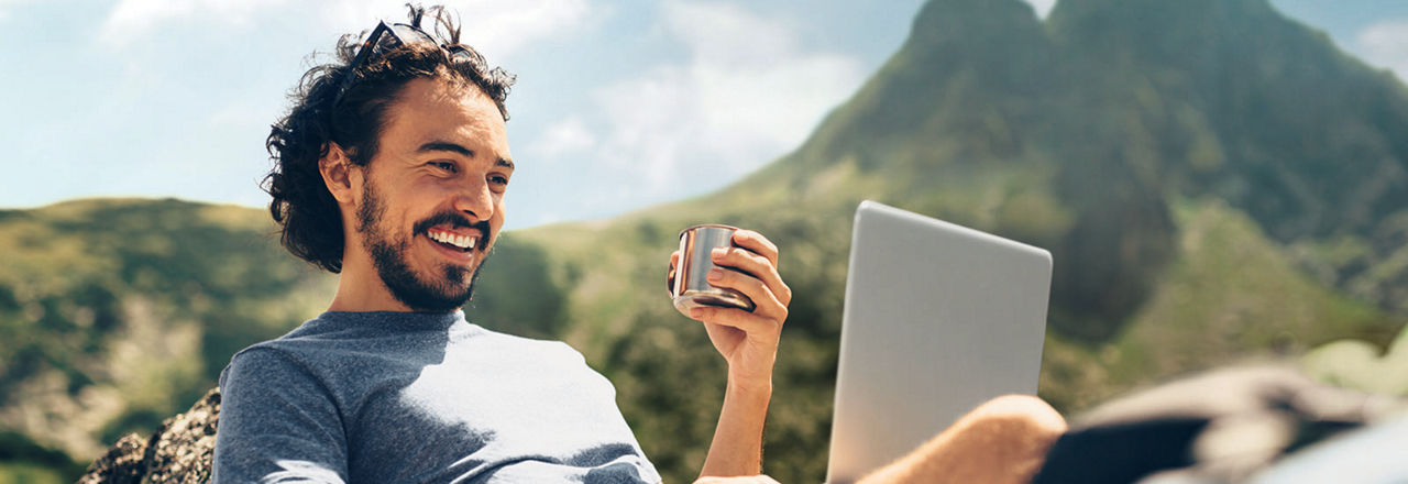 Male hiker having a drink while he's using a laptop on a rock near a lake in the mountain.