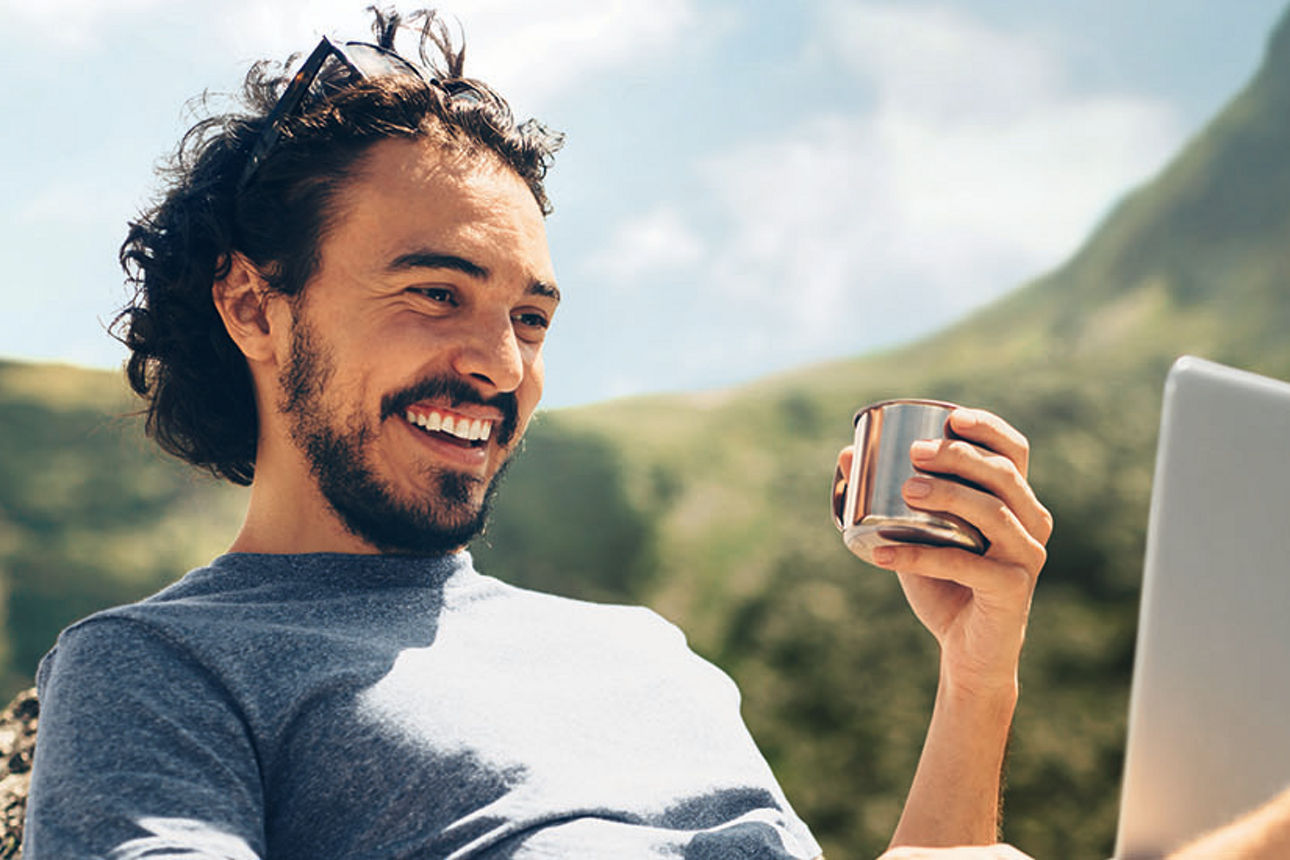 Male hiker having a drink while he's using a laptop on a rock near a lake in the mountain.
