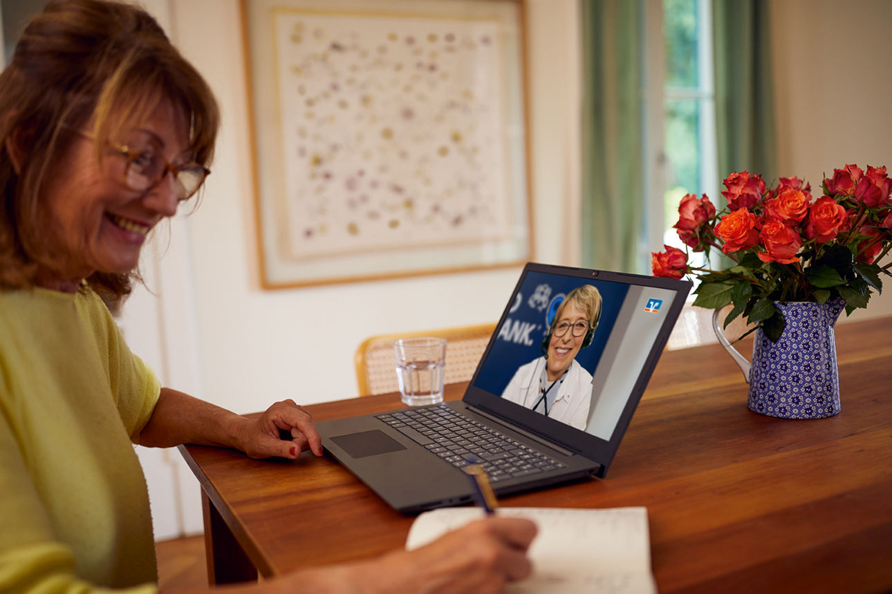 Ältere Frau sitzt an einem Holztisch und macht Notizen während einer Video-Beratung auf einem Laptop