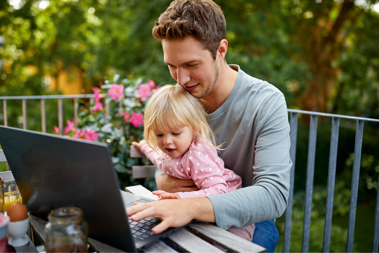 Junger Mann mit Mädchen auf dem Arm sitzen am Tisch und arbeiten am Laptop