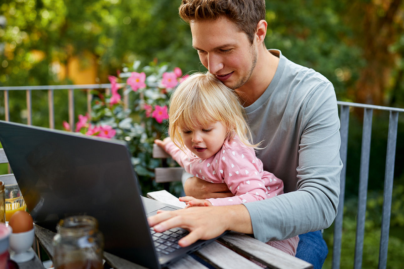 Junger Vater sitzt mit seiner Tochter auf dem Schoß vor einem Laptop.