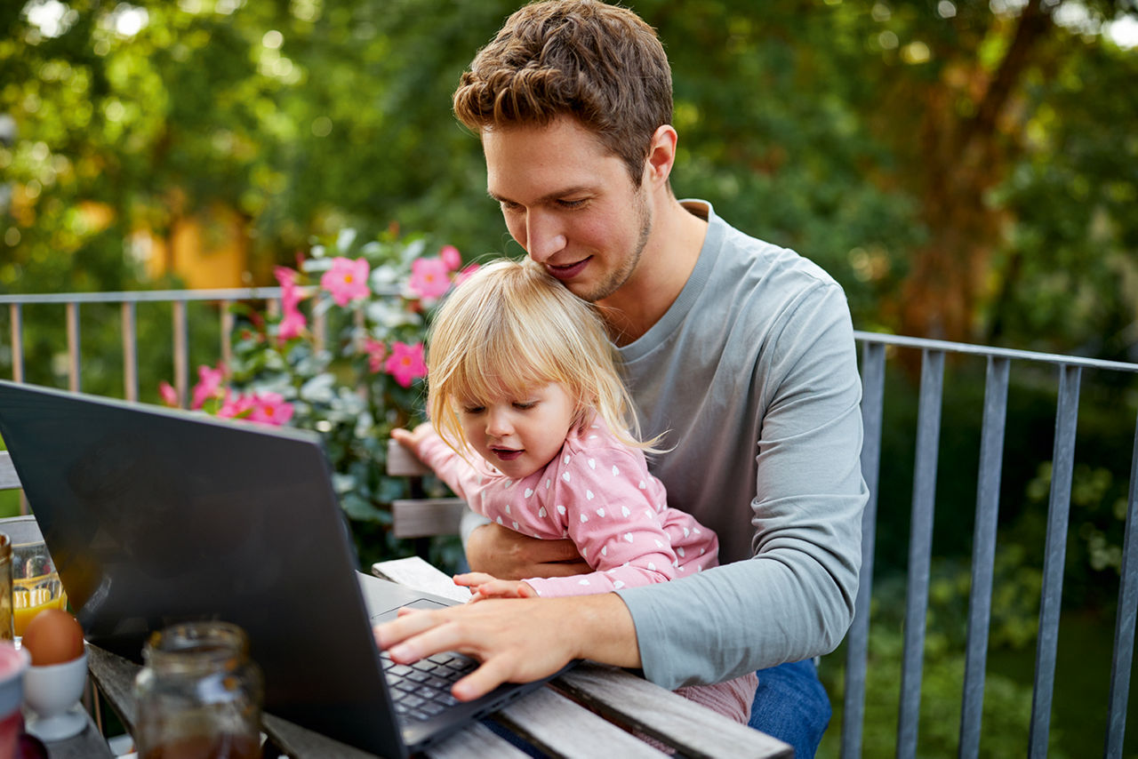 Ein Vater arbeitet auf dem Laptop auf einem Balkon, während seine kleine Tochter lachend auf seinem Arm sitzt