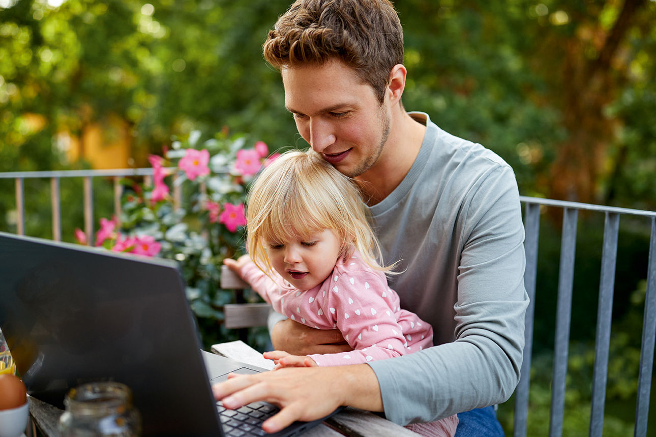 Ein Vater arbeitet auf dem Laptop auf einem Balkon, während seine kleine Tochter lachend auf seinem Arm sitzt