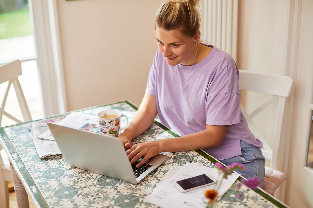 Frau arbeitet entspannt am Laptop am Küchentisch, umgeben von Unterlagen, Handy und einer Tasse Tee.