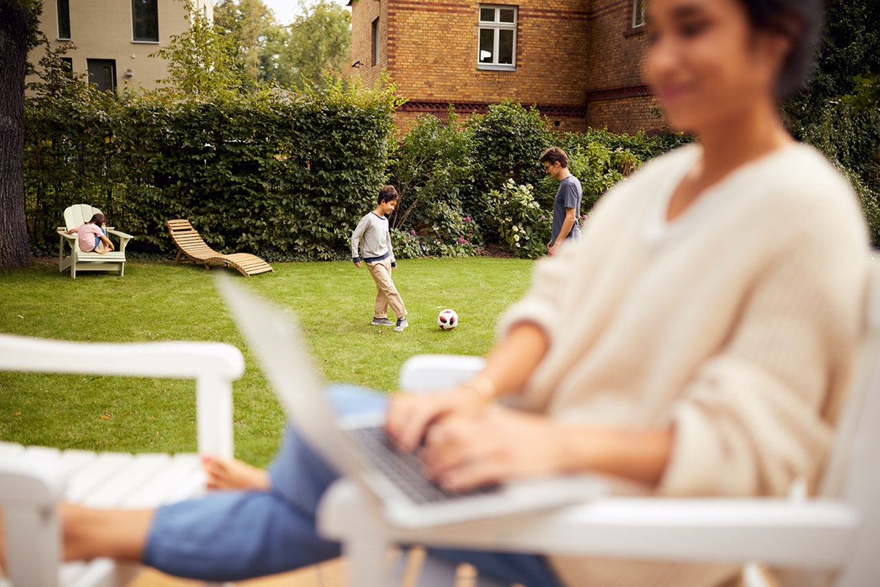 Frau sitzt im GArten am Laptop und schaut der Familie zu