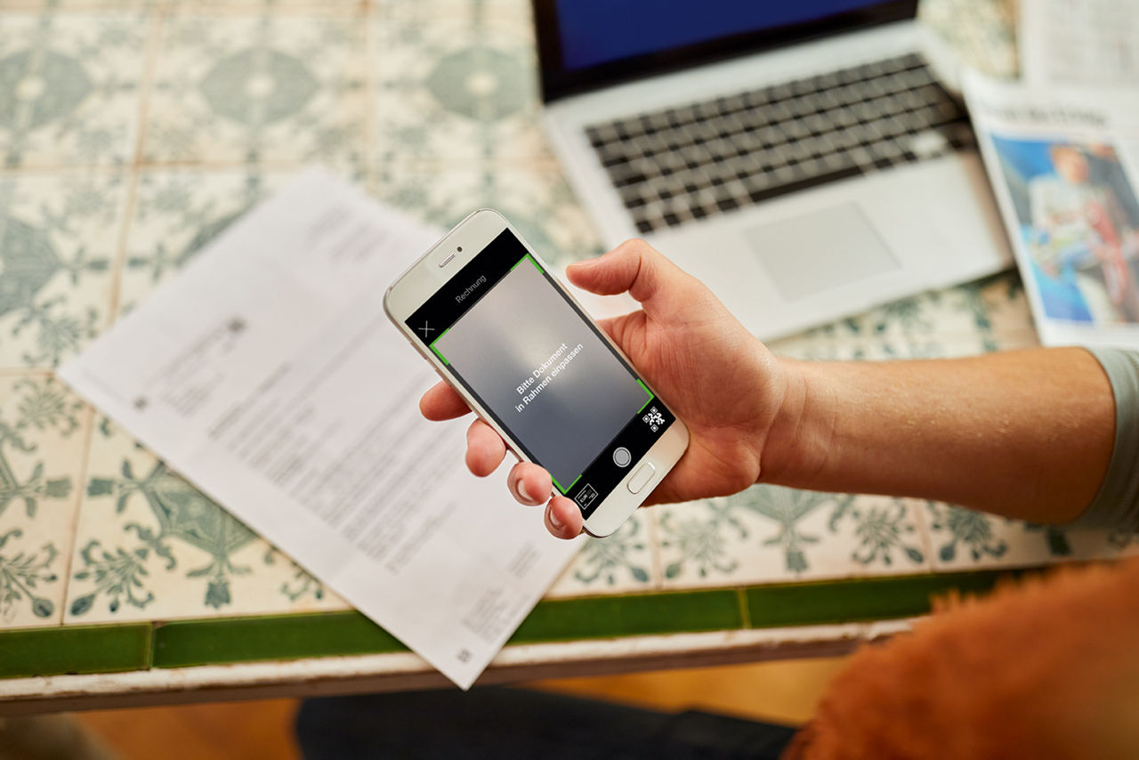 Person hält Handy, im Hintergrund ist ein Brief und eine Laptop zu sehen