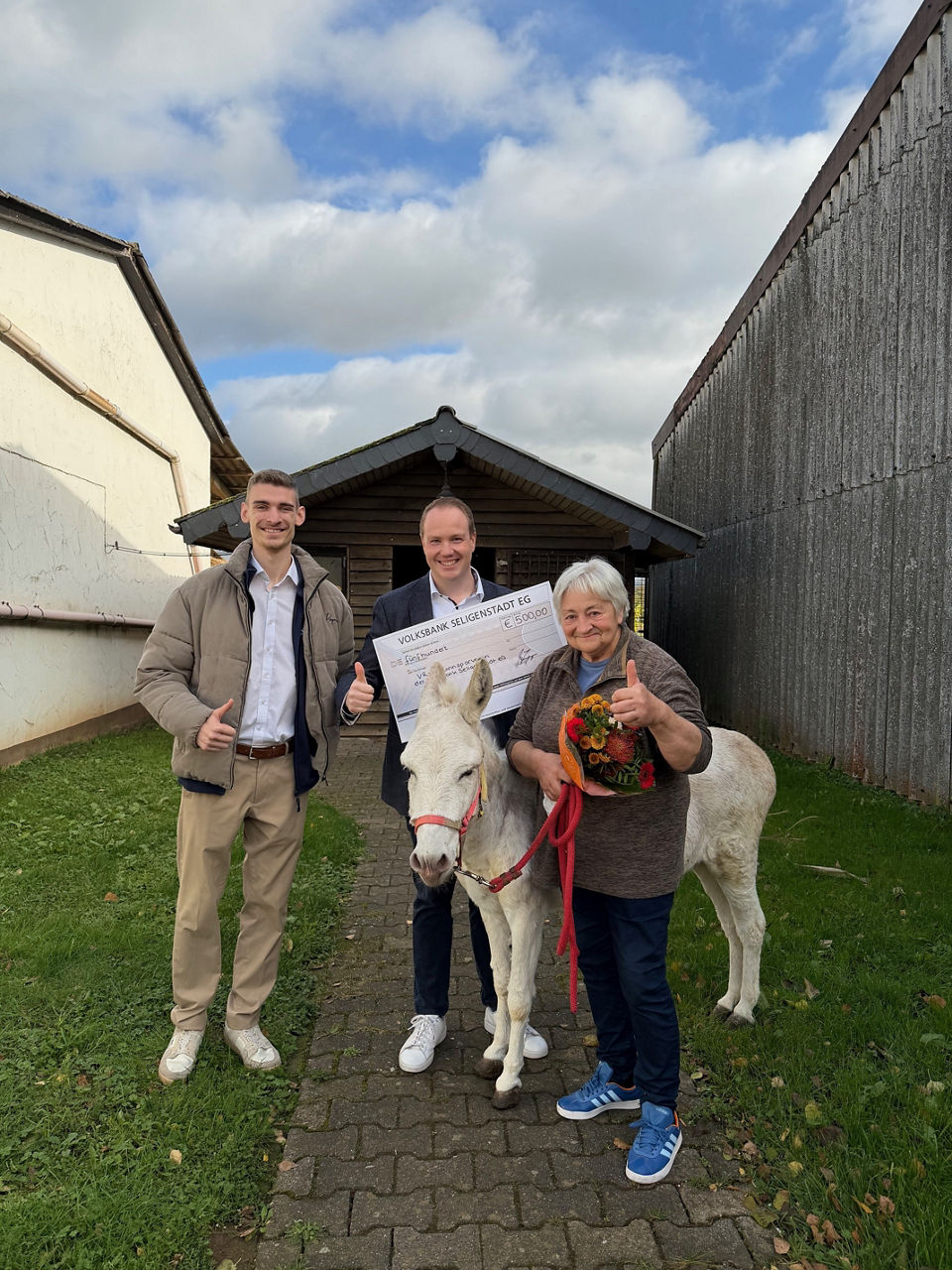 Zwei Männer und eine Frau posieren im Freien und halten einen großen Scheck in den Händen.