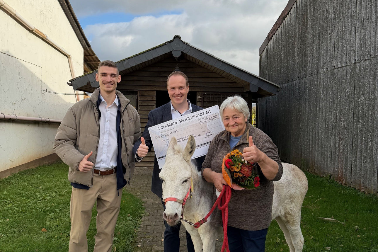 Zwei Männer und eine Frau posieren im Freien und halten einen großen Scheck in den Händen.