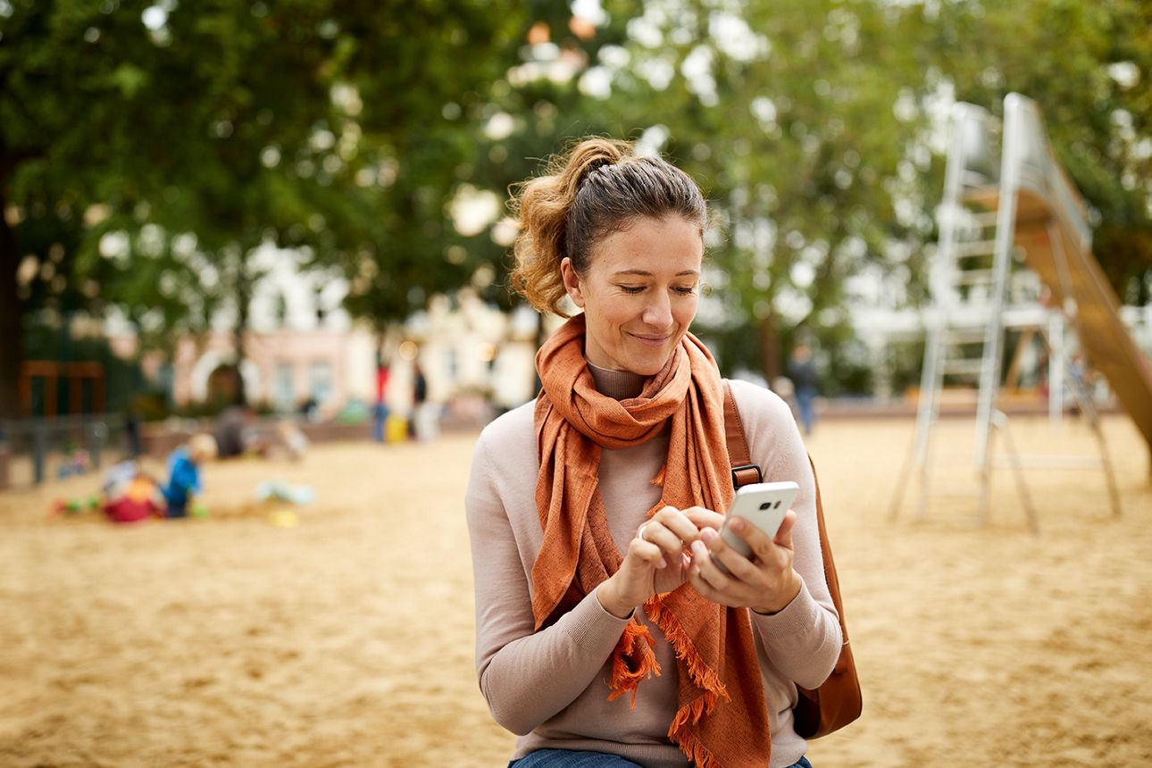 Eine Frau steht auf einem Spielplatz und tippt auf das Display ihres Handys, das sie in der Hand hält.