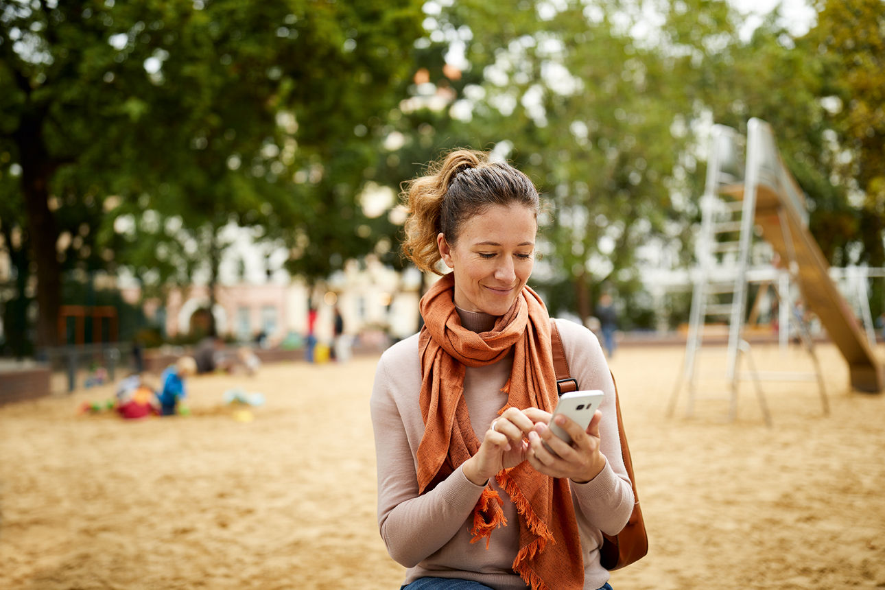 Frau auf einem Spielplatz checkt Ihr Girokonto bequem auf dem Handy