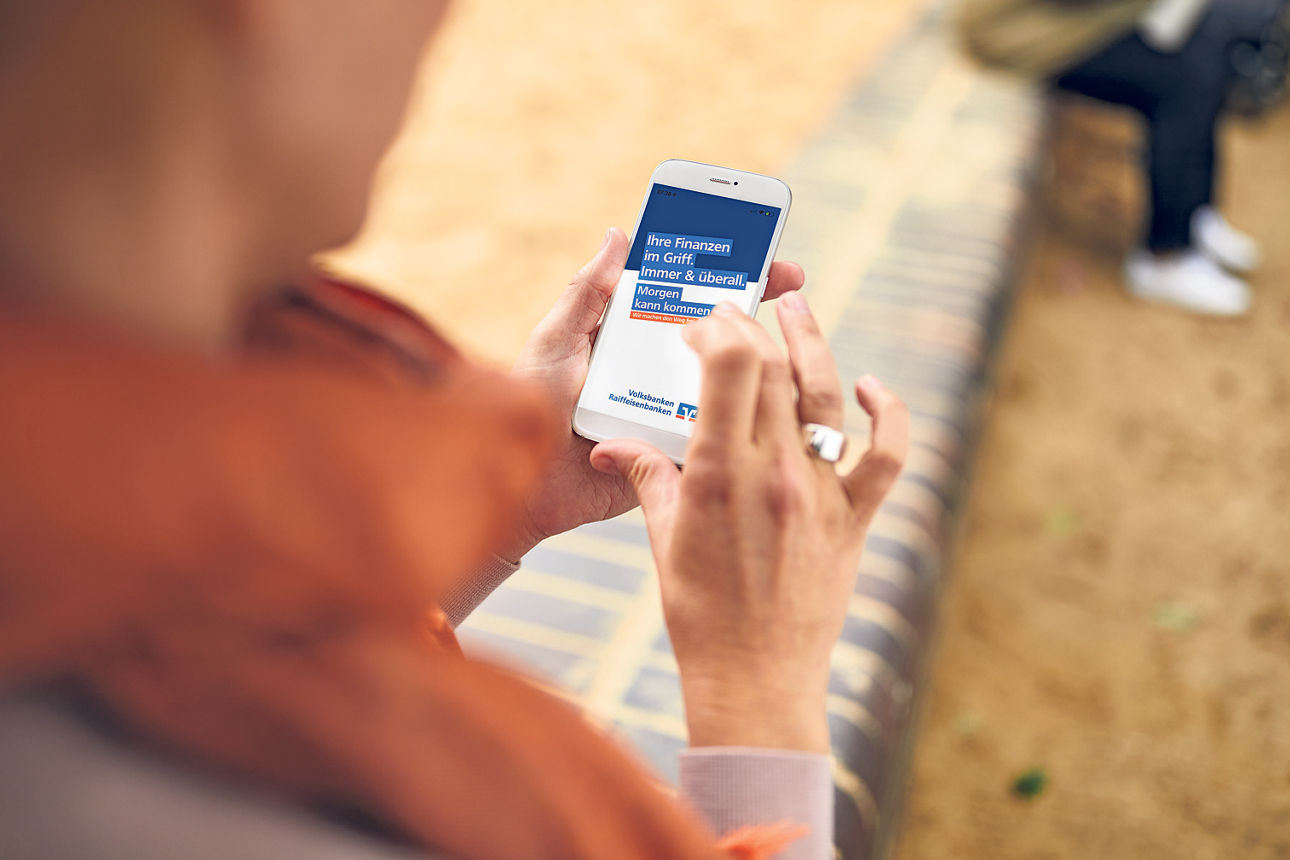Eine Person hält ein Smartphone mit einer App in der Hand, auf der der Text „Ihre Finanzen im Griff: Immer & überall“ angezei