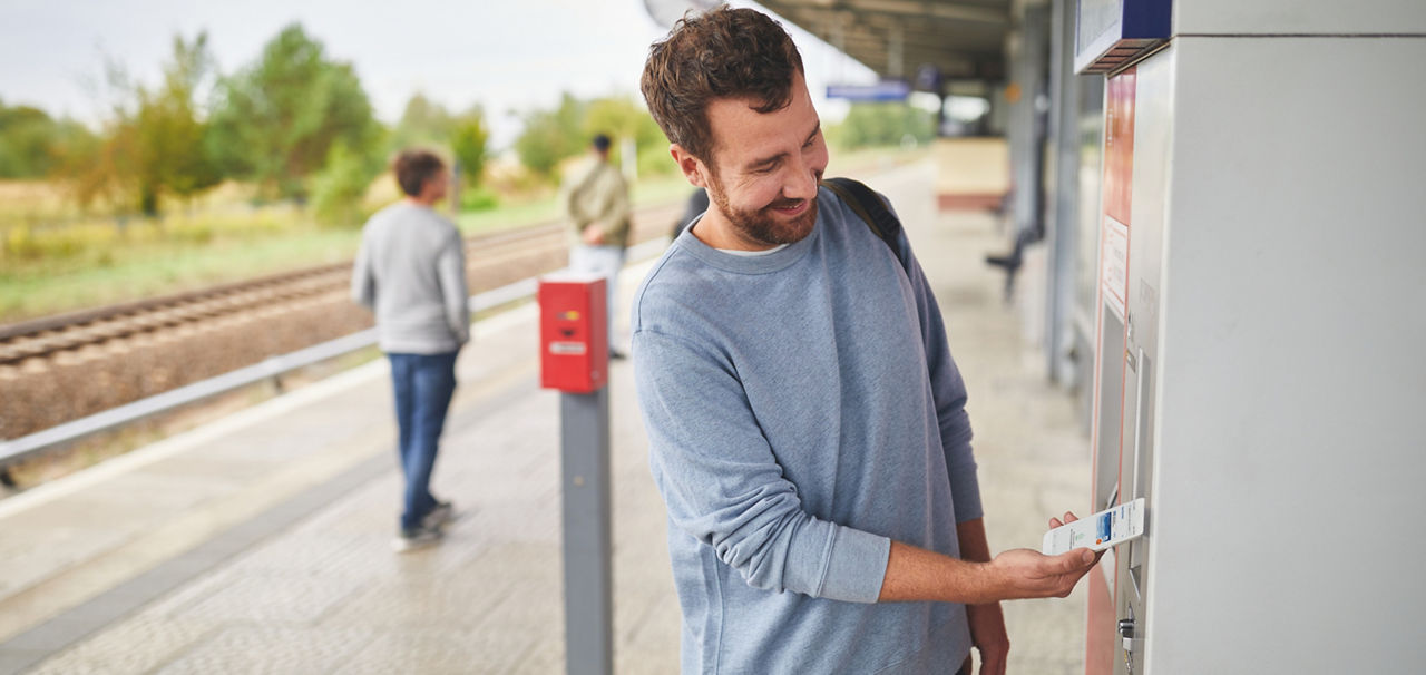 Mann bezahlt auf einem Bahnsteig am Ticketautomaten mit dem Handy 