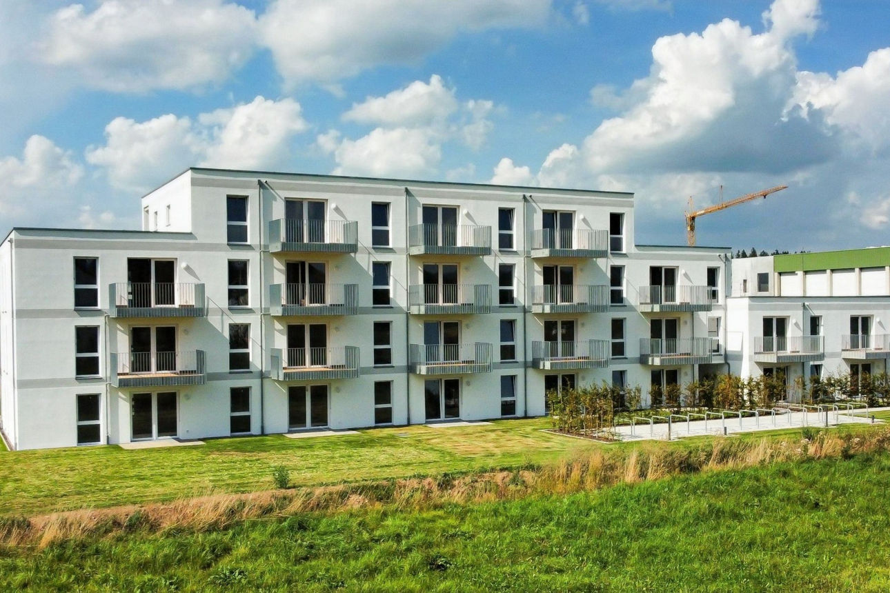 Weißes Neubau Mehrfamilienhaus. Davor eine grüne Wiese. Der Himmel ist blau.