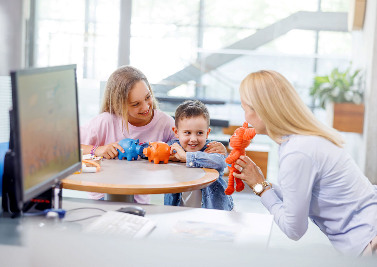 Eine Bankmitarbeiterin zeigt zwei Kindern mit Ihren Spardosen ein Kuscheltier in orange.