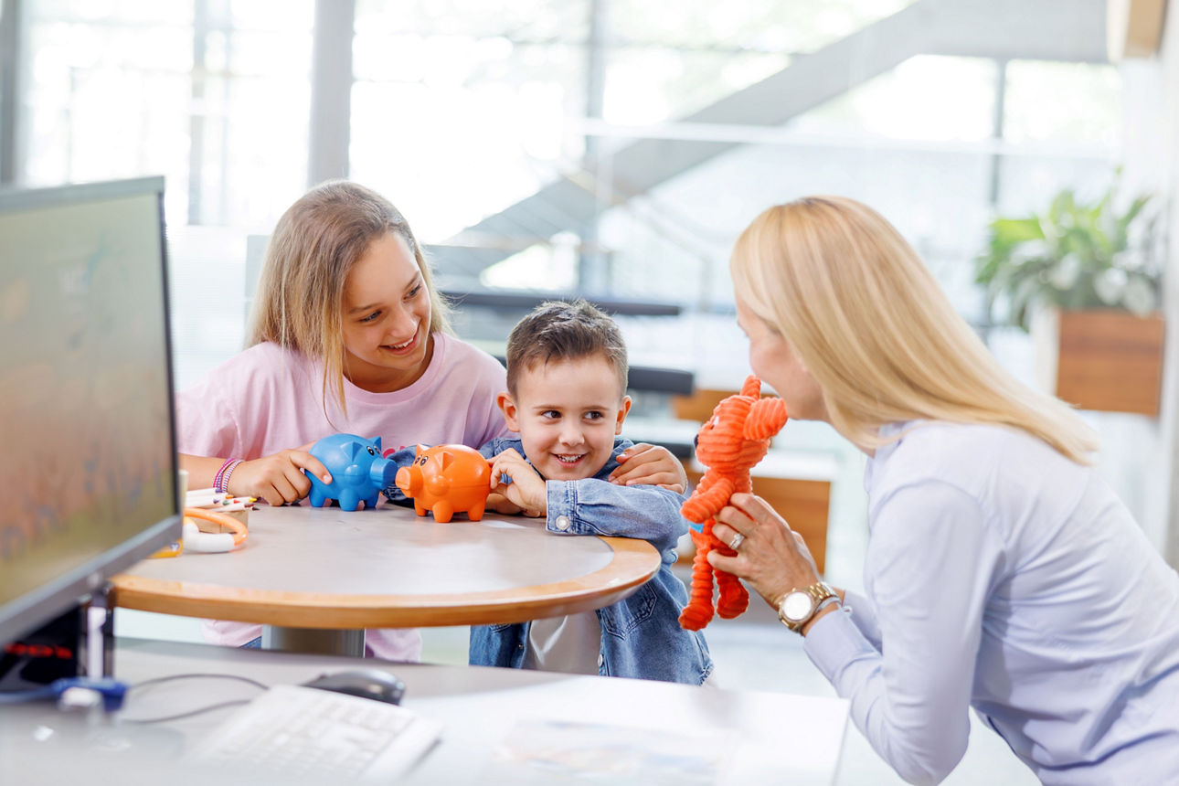 Eine Bankmitarbeiterin zeigt zwei Kindern mit Ihren Spardosen ein Kuscheltier in orange.