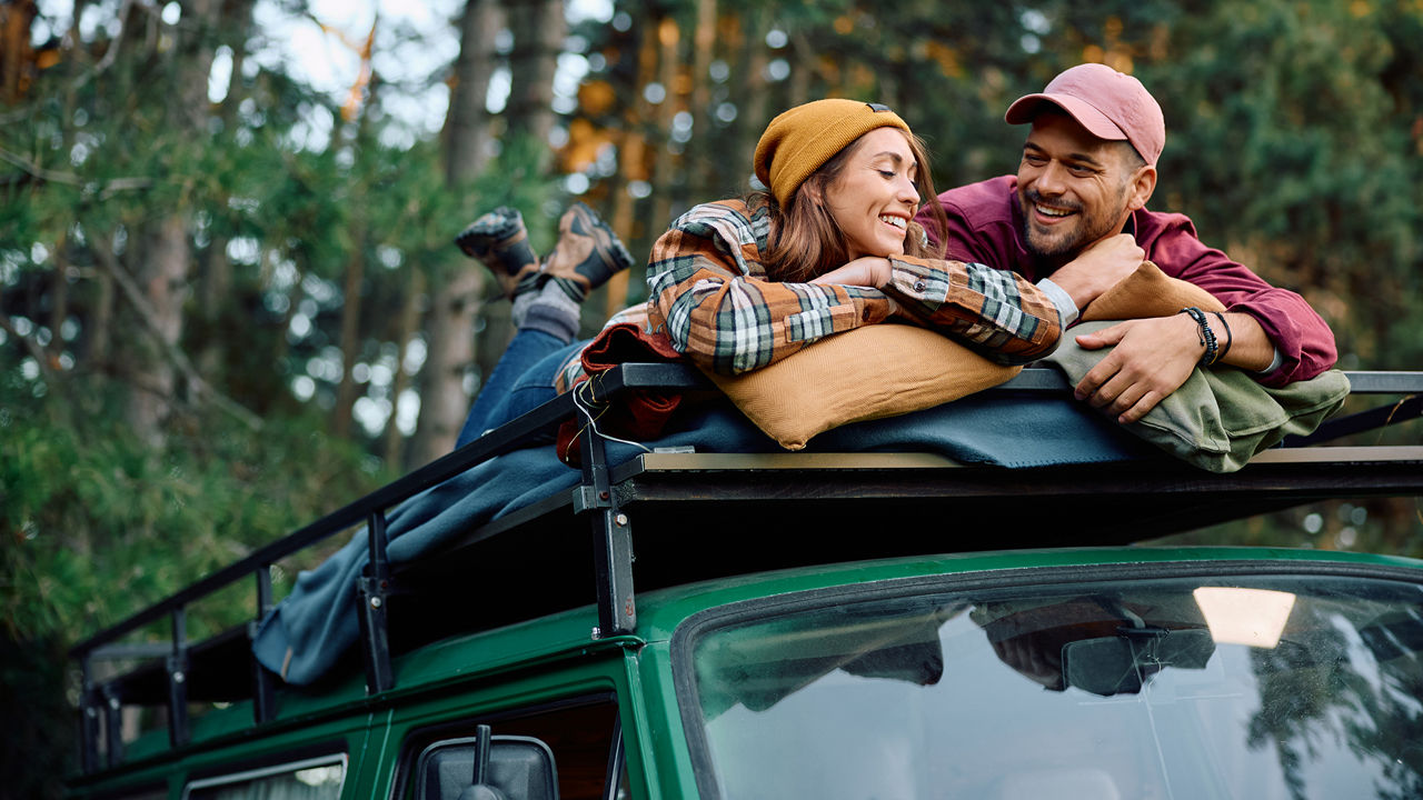 Young happy couple communicating while relaxing on camper trailer's roof in nature