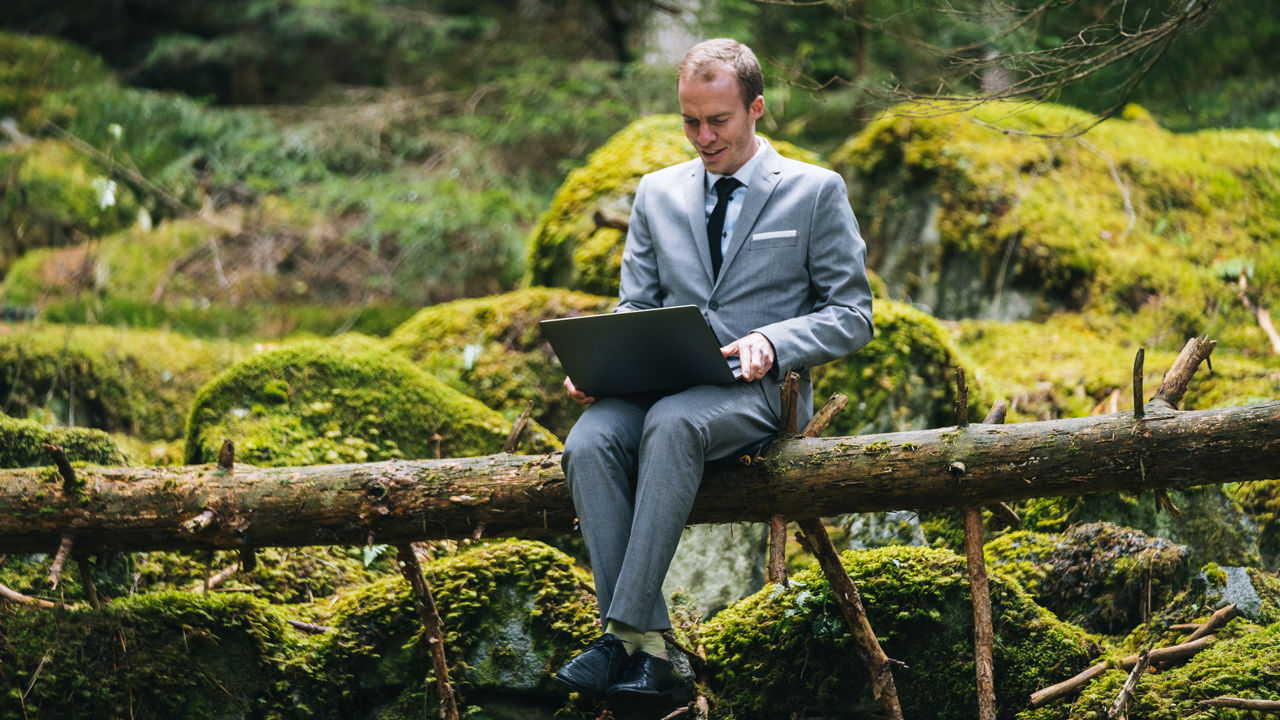 Ein Mann trägt einen Anzug und sitzt im Wald auf einem Baumstamm und arbeitet an seinem Laptop