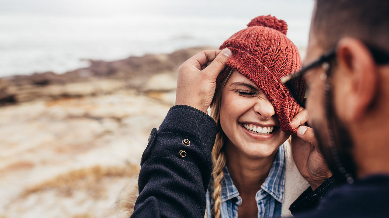 Ein Mann zieht am Strand einer jungen Frau ihre orange Mütze über ihr linkes Auge. Die Frau lacht. 