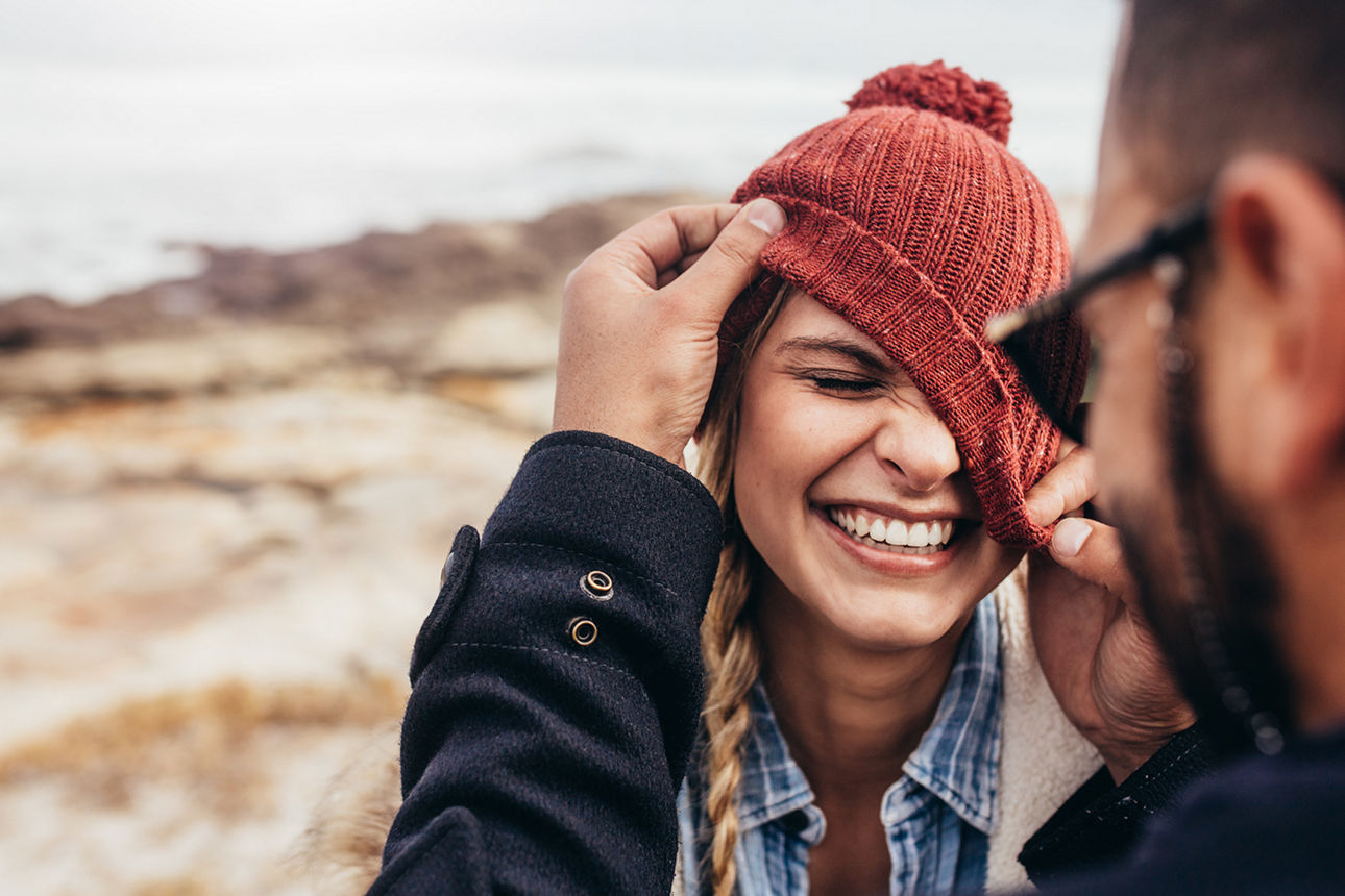 Ein Mann zieht am Strand einer jungen Frau ihre orange Mütze über ihr linkes Auge. Die Frau lacht. 