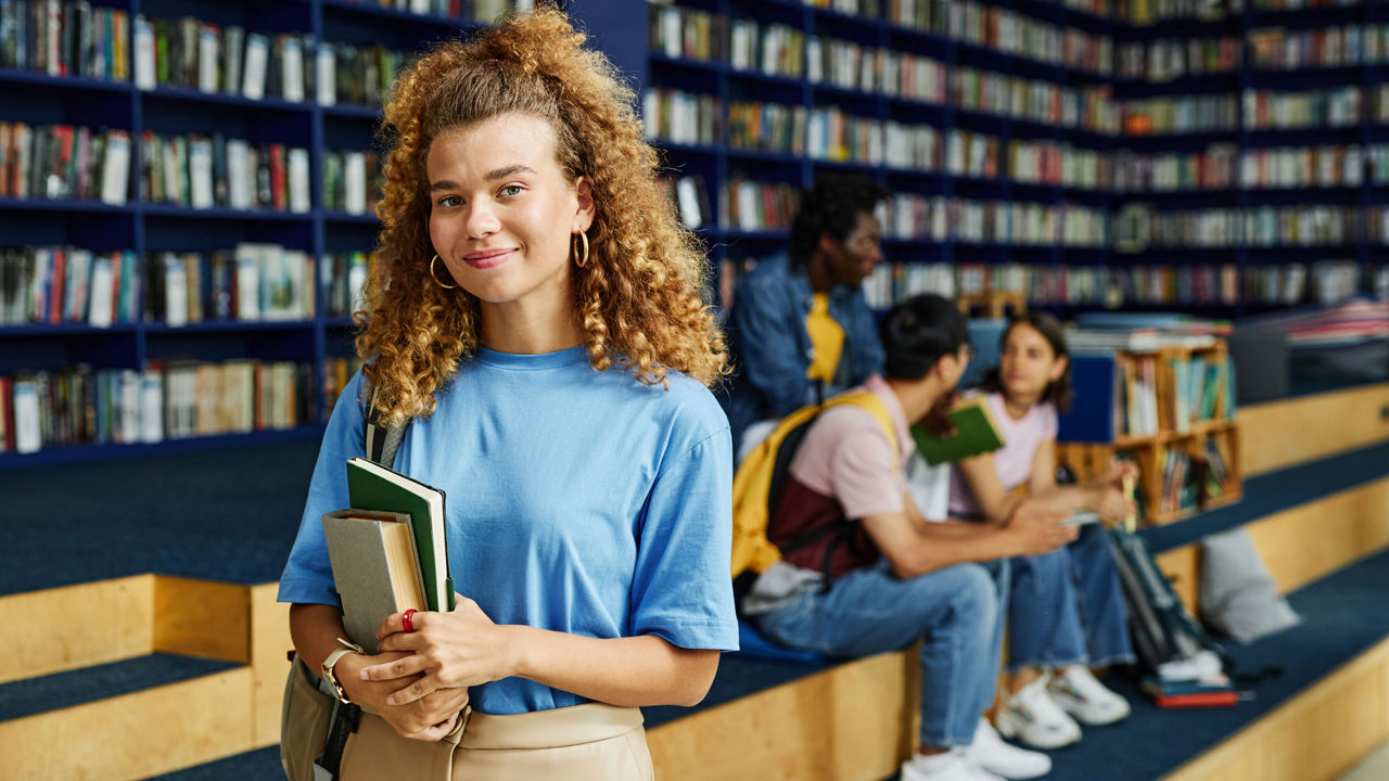 Im Vordergrund steht eine Studentin und lächelt in die Kamera. Hinter ihr ist eine Bücherwand und drei sitzende Studenten.