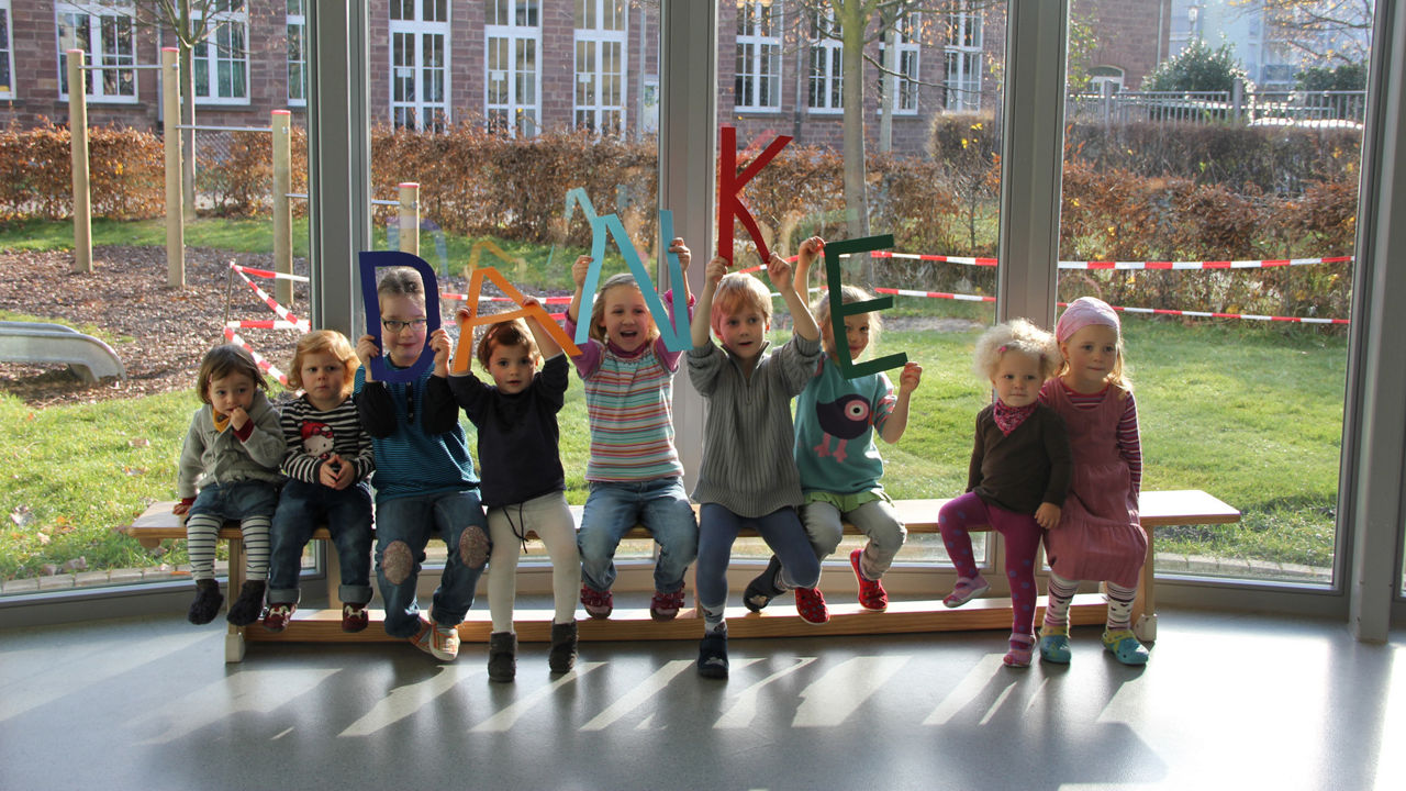 Neun Kinder, die nebeneinander sitzen und Buchstaben hoch halten, die das Wort Danke formen.