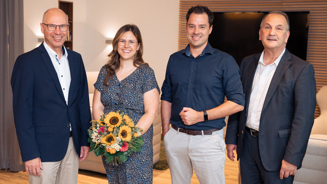 Vier Personen, die lächelnd in die Kamera schauen. Der Frau im Foto hat ein Stipendium und einen Blumenstrauß erhalten.