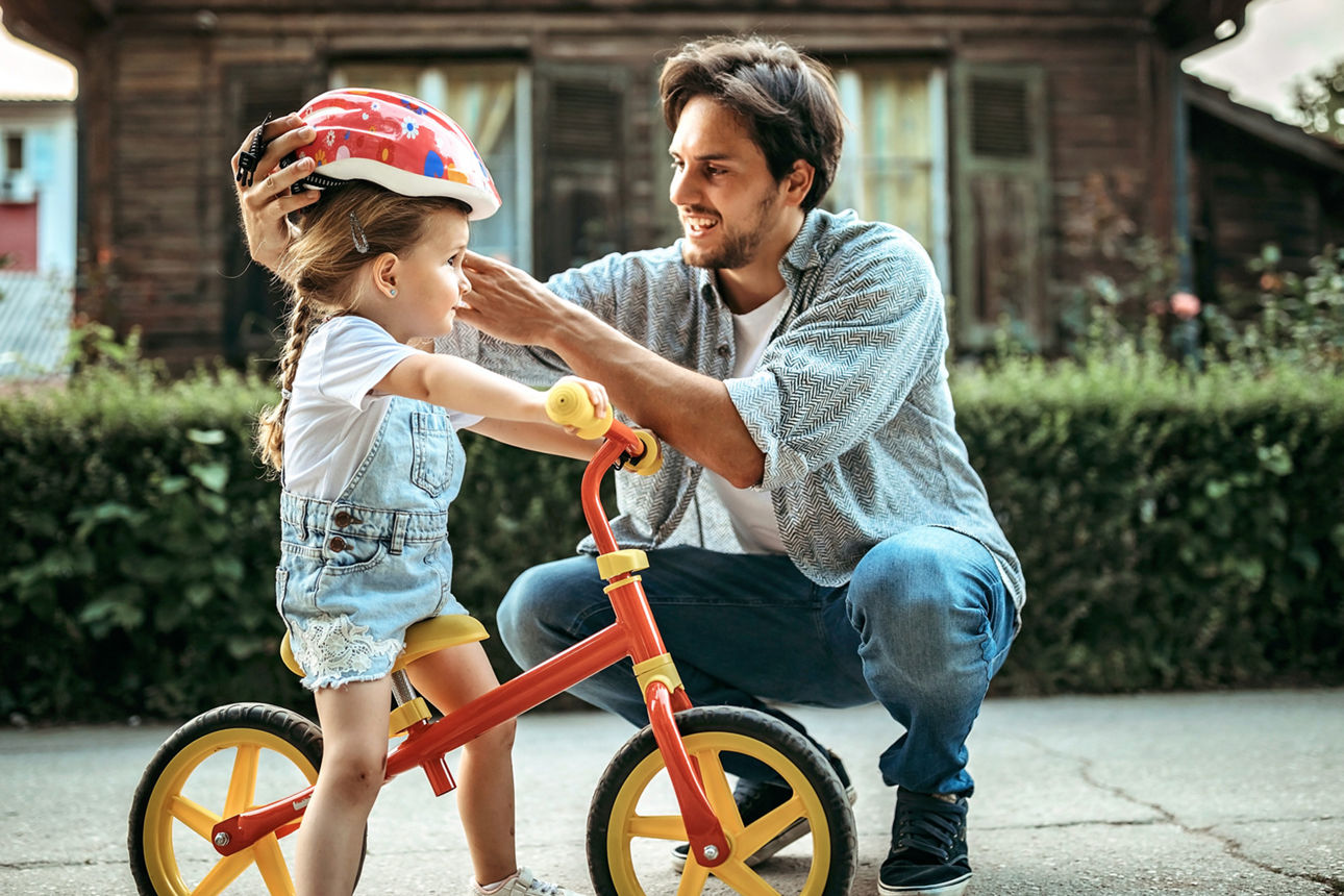 Ein Mann setzt einem kleinen Mädchen auf einem Laufrad einen Helm auf. 