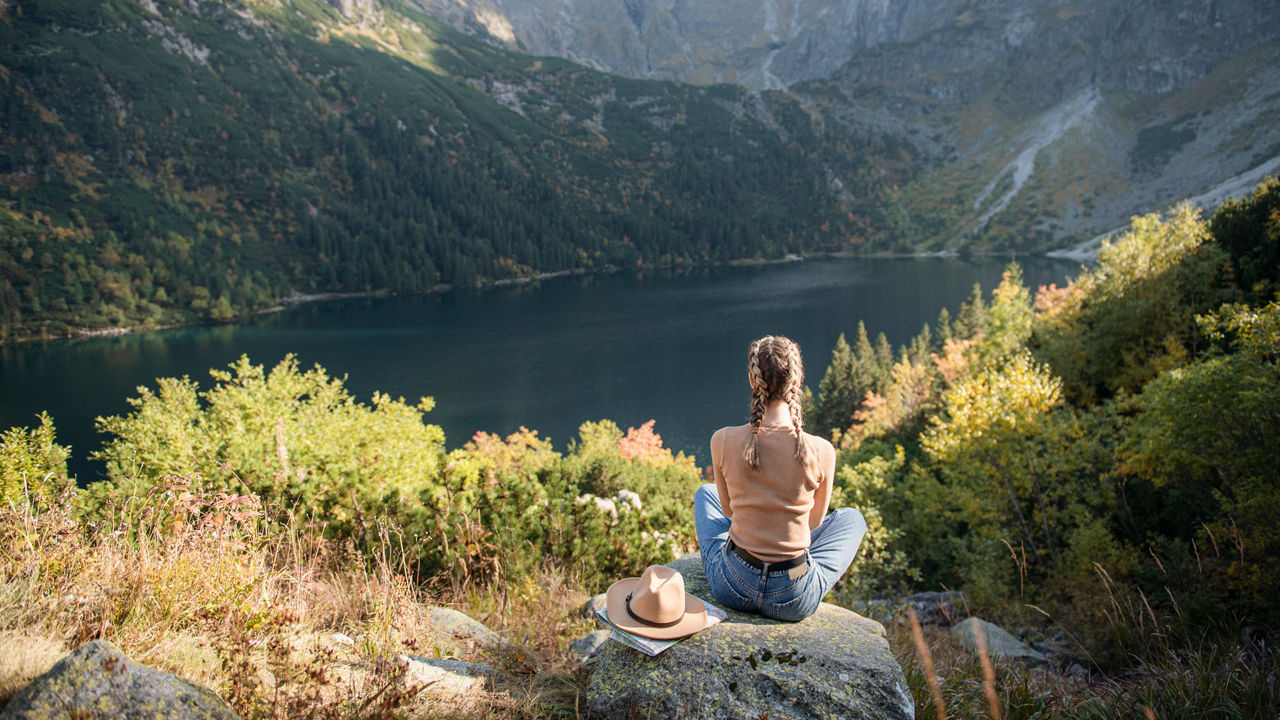 Eine bergige Landschaft mit einem See in der Mitte. Unten rechts sitzt eine Frau im Schneidesrsitz auf einem Stein.
