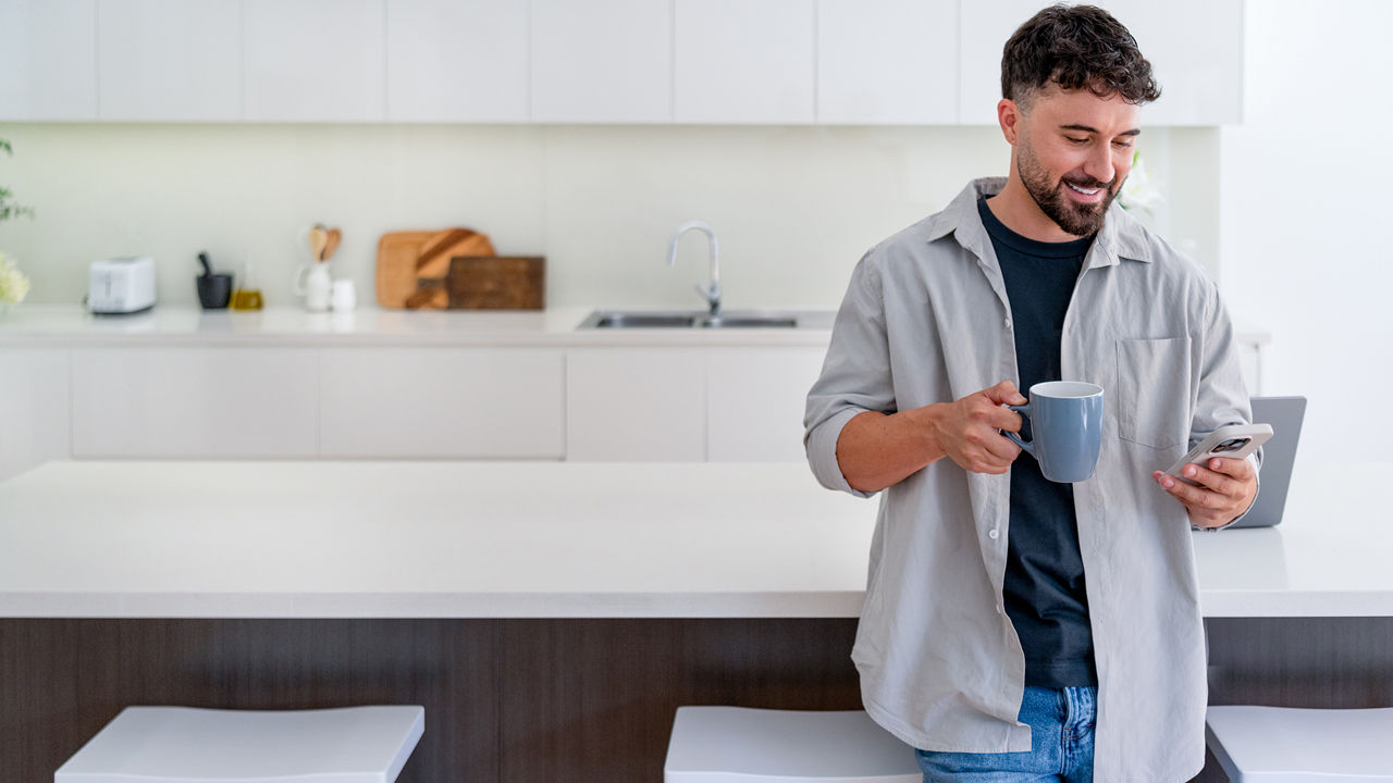 Ein Mann steht in der Küche, trinkt Kaffee und schaut lächelnd in sein Handy.